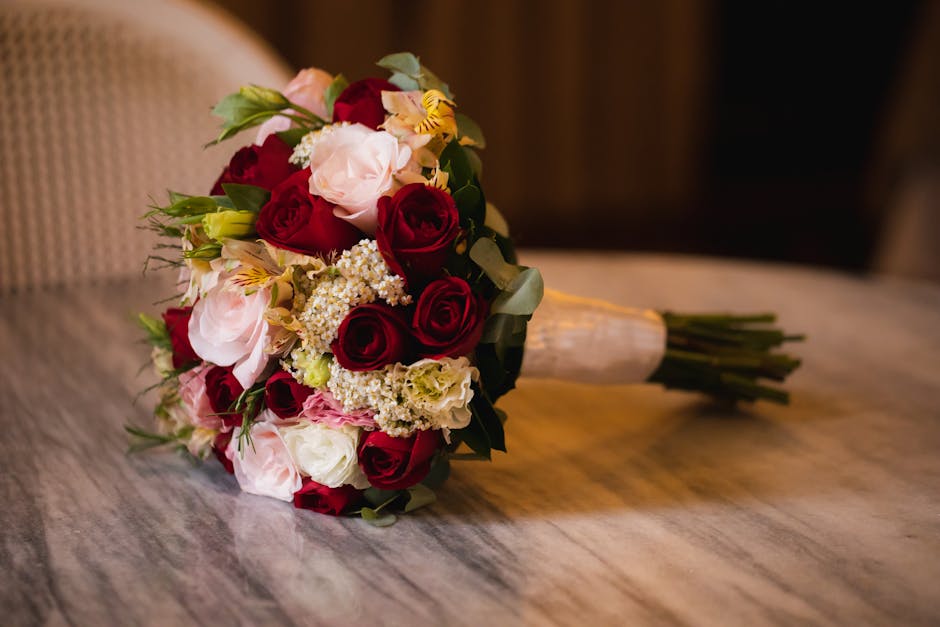 A bouquet of fresh flowers featuring deep red roses, pale pink roses, white roses, and small clusters of white baby's breath, arranged with green foliage. The bouquet is wrapped with light-colored pap