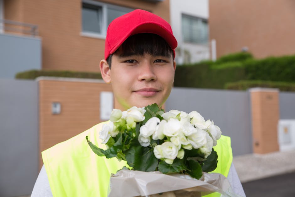 A young male delivery worker with light skin, dark hair, and a gentle smile, wearing a red cap and a high-visibility yellow vest, holding a bouquet of fresh white roses wrapped in white paper. The bac