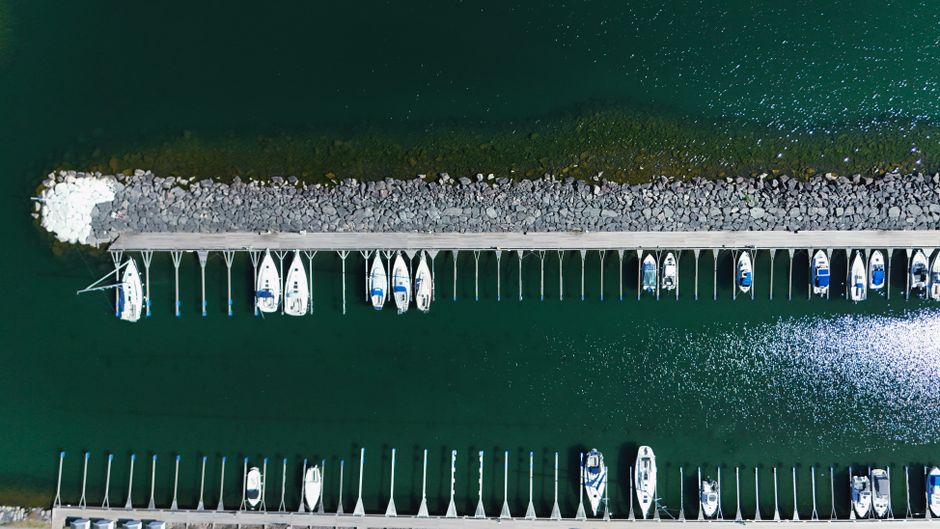 Aerial view of a marina with a long, narrow pier extending into calm, dark green water, lined with evenly spaced white supports with blue and white boats docked on either side. The boats vary in size