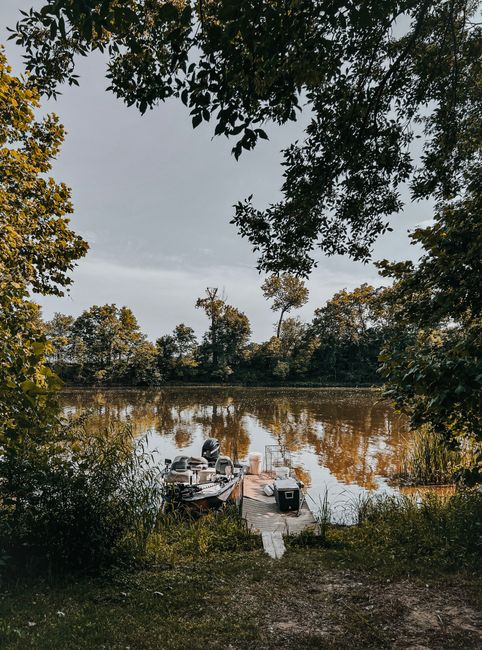A serene lakeside scene featuring lush green trees framing the view, with a small wooden dock extending into calm, reflective water. On the dock, there is a boat with visible steering equipment and se