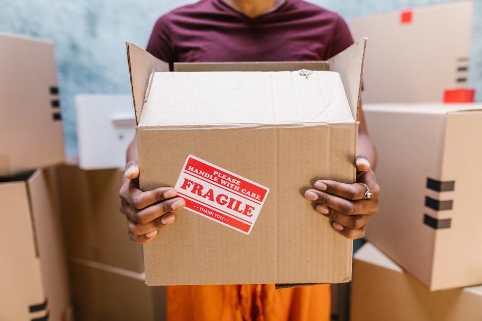 A person holding an open cardboard shipping box with a red and white 'Fragile Handle with Care' sticker on the front. The individual is standing among several closed cardboard boxes, suggesting a pack